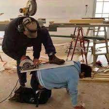 Foto de un hombre usando a otro como soporte para cortar una tabla con una sierra circular.